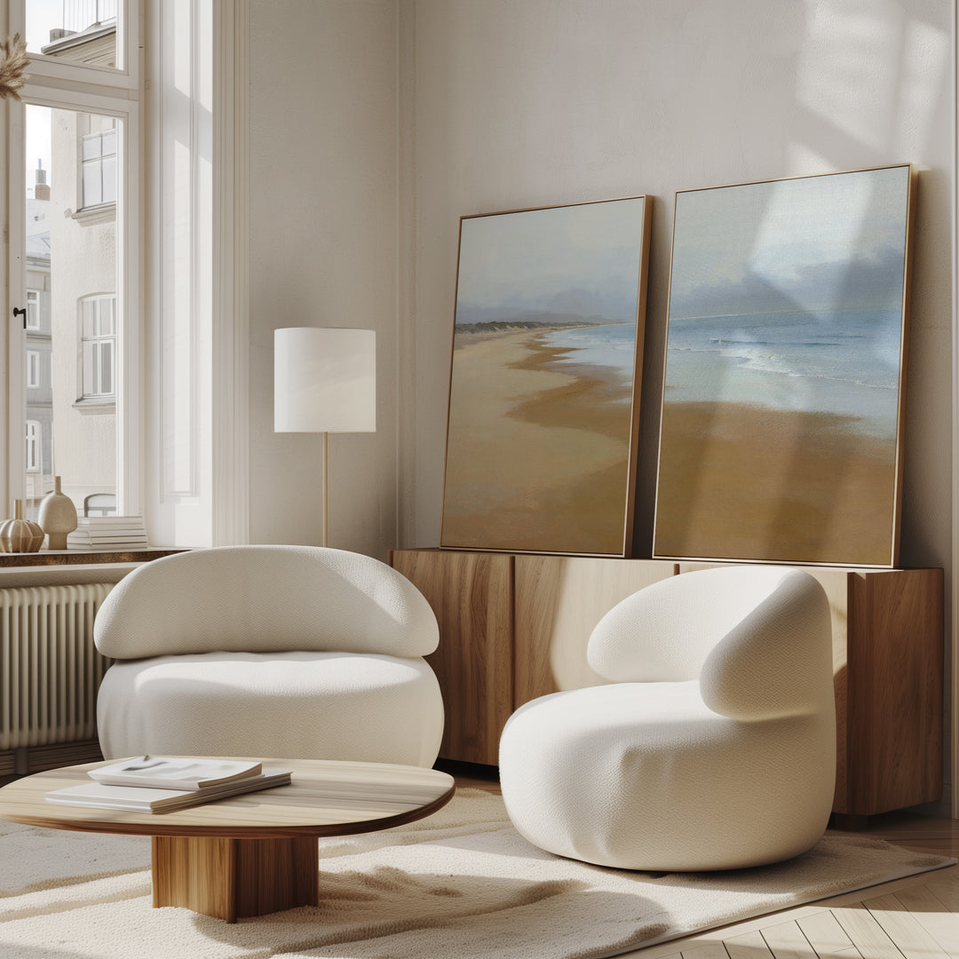 Two modern white curved armchairs and a low wooden table sit in a bright living room. The "Endless Shoreline - Set of 2" wall art rests against the wall on a wooden cabinet, enhancing the tranquil atmosphere with its beach scenes.