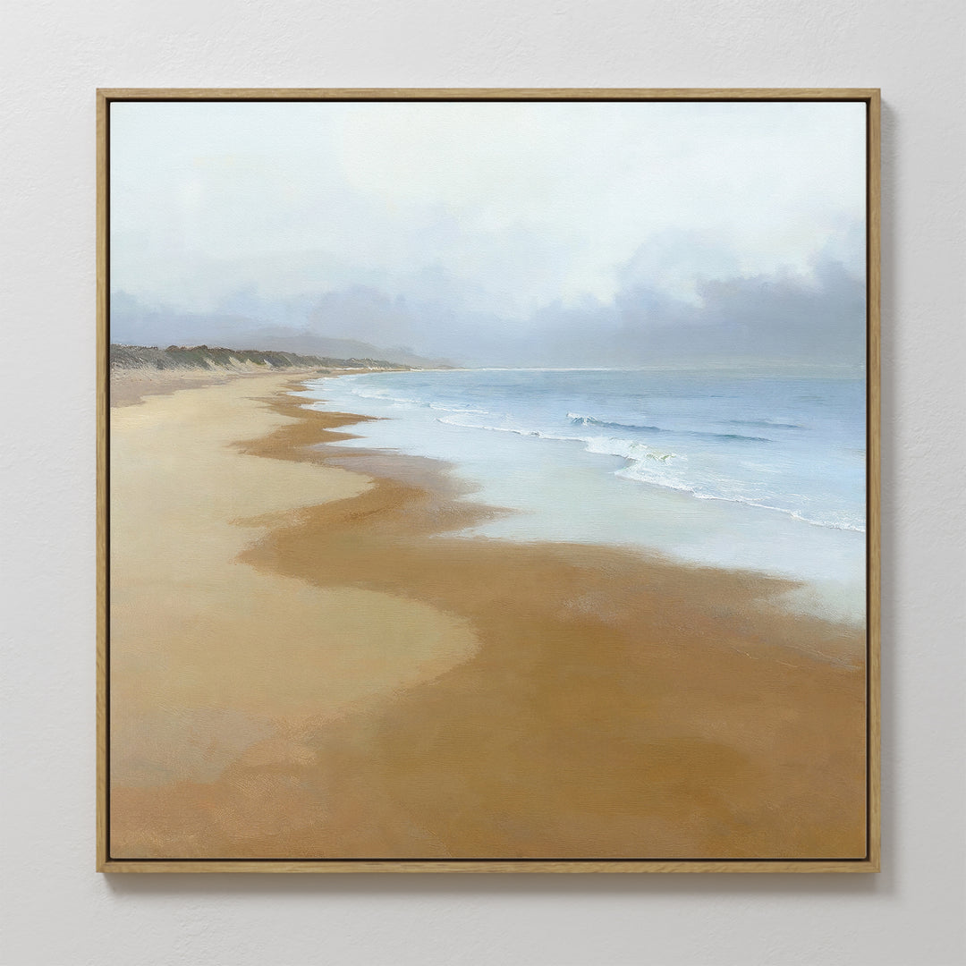 The Endless Shoreline Canvas Art - Square, featuring a peaceful beach with gentle waves, light brown sand, and a cloudy sky, hangs on a white wall for a serene coastal touch.