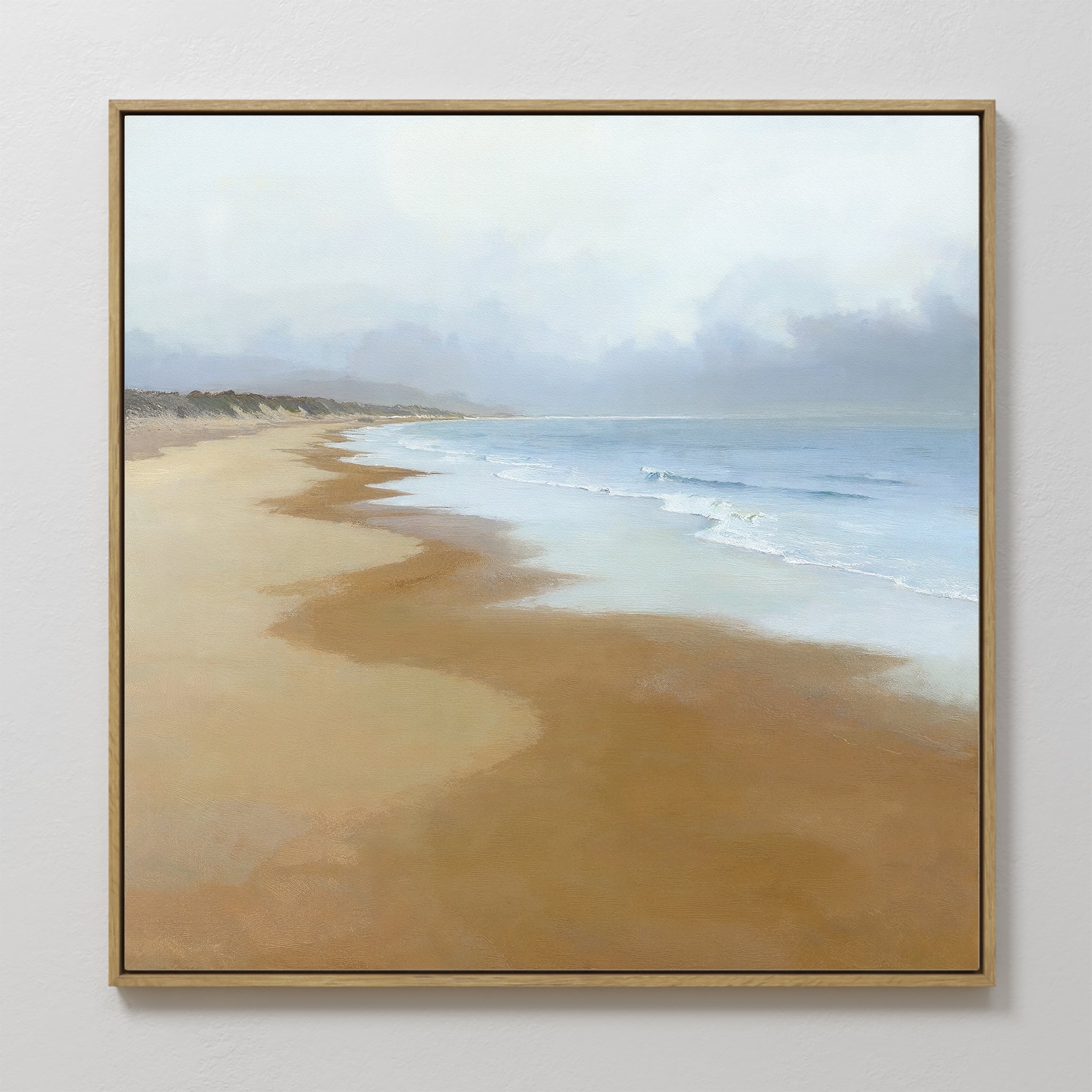 The Endless Shoreline Canvas Art - Square, featuring a peaceful beach with gentle waves, light brown sand, and a cloudy sky, hangs on a white wall for a serene coastal touch.