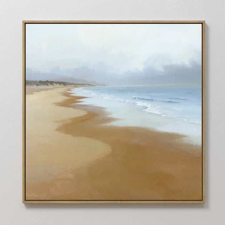 The Endless Shoreline Canvas Art - Square, featuring a peaceful beach with gentle waves, light brown sand, and a cloudy sky, hangs on a white wall for a serene coastal touch.