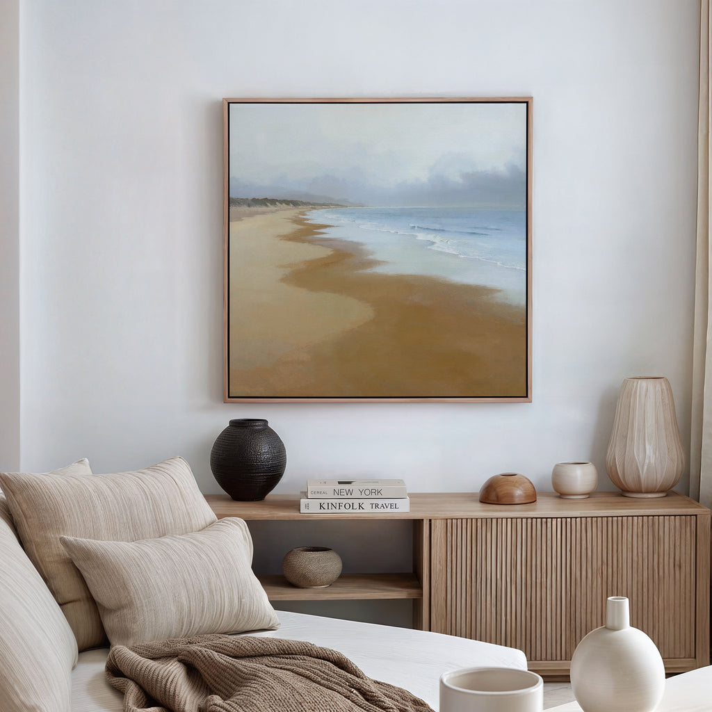 A minimalist living room features a beige sofa with cushions, a wooden sideboard with vases and books, and the Endless Shoreline Canvas Art - Square as large framed wall art on a white wall. Natural light enters from the right.