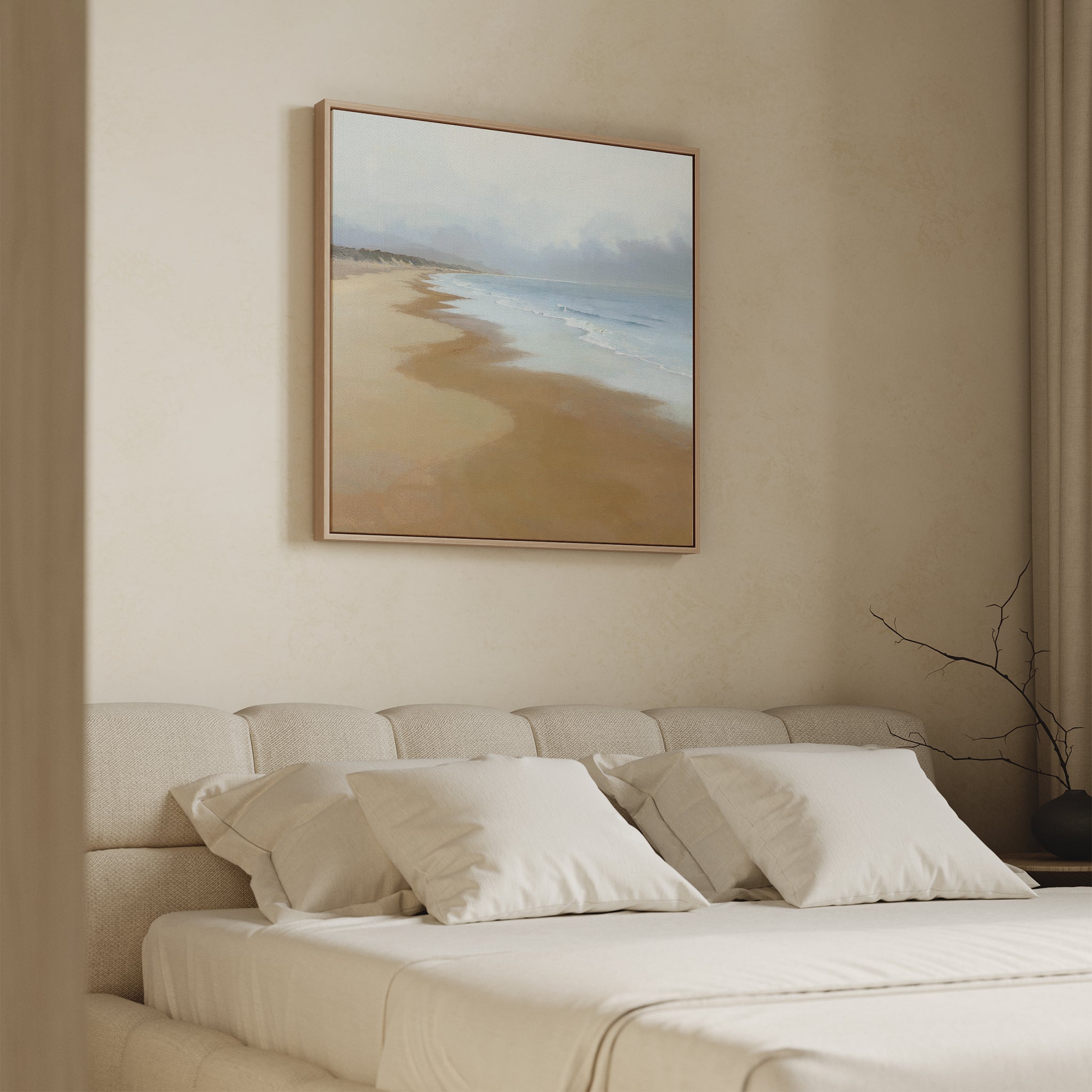 A beige, minimalist bedroom features a low tufted headboard with tidy pillows and the Endless Shoreline Canvas Art - Square above the bed, showcasing a serene coastal scene of sand, sea, and clouds.