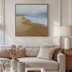 A cozy living room features a beige sofa, neutral pillows, a side table with a lamp, and a vase of dried flowers. Above the sofa hangs the Endless Shoreline Canvas Art - Square, showcasing a tranquil coastal scene.