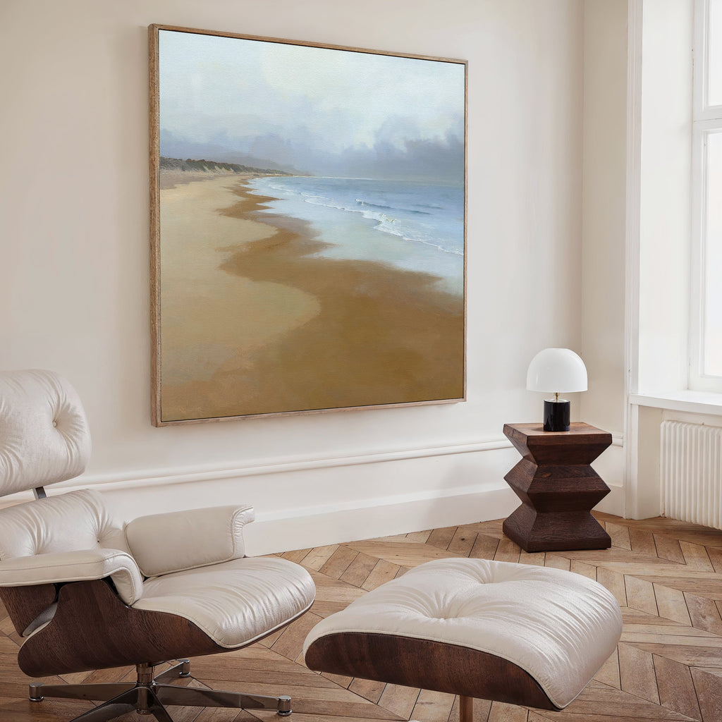A modern living room featuring a leather lounge chair and ottoman, a wooden side table with lamp, and the Endless Shoreline Canvas Art - Square—a large coastal print—hanging on a beige wall for serene decor.