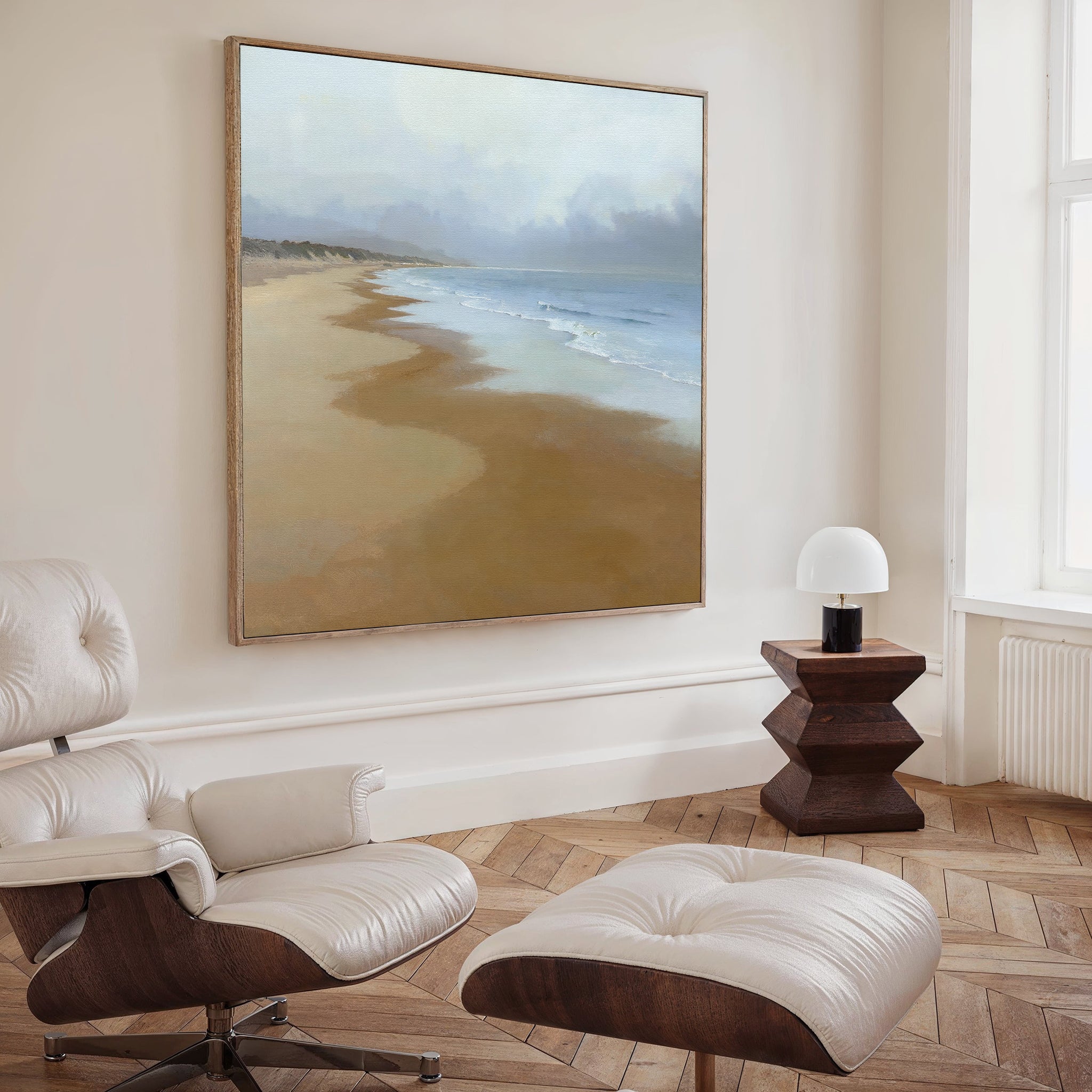 A modern living room featuring a leather lounge chair and ottoman, a wooden side table with lamp, and the Endless Shoreline Canvas Art - Square—a large coastal print—hanging on a beige wall for serene decor.