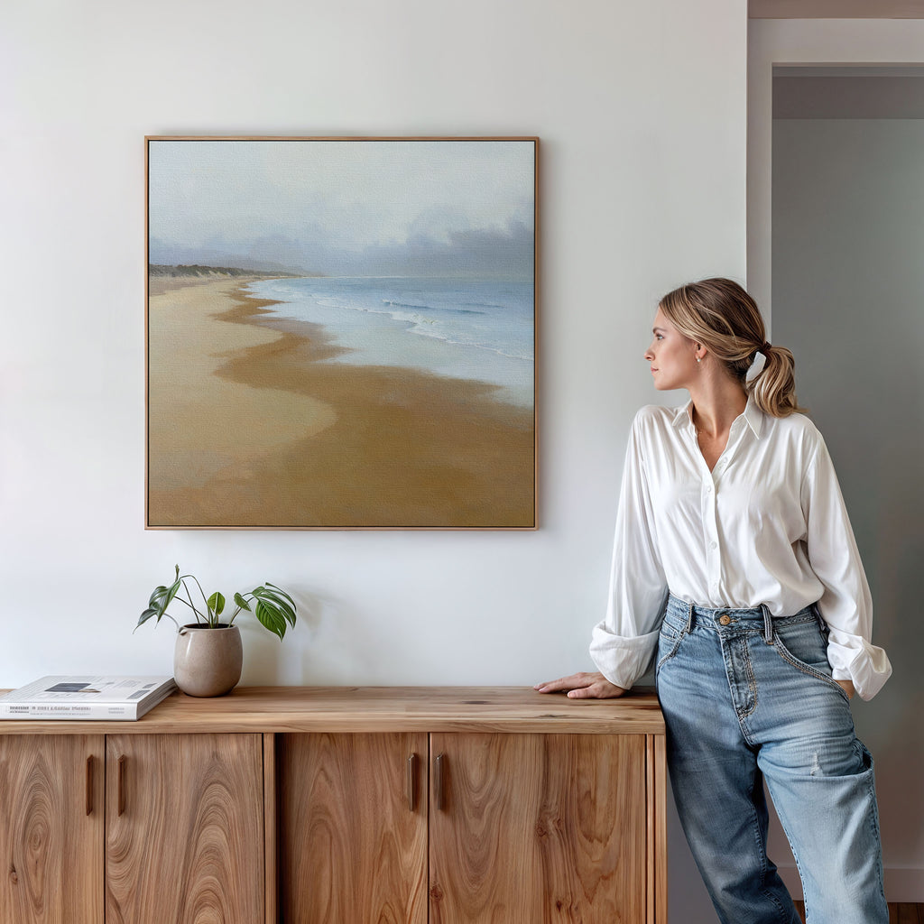 A woman in a white blouse and jeans admires the Endless Shoreline Canvas Art - Square above a wooden cabinet. A potted plant and book add to the serene, modern, minimalistic decor of the room.