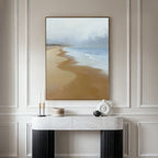 The Endless Shoreline Canvas Art - Vertical, featuring a serene coastal scene, hangs on a beige wall above a marble console table with black legs, adorned with decorative candles and vases.