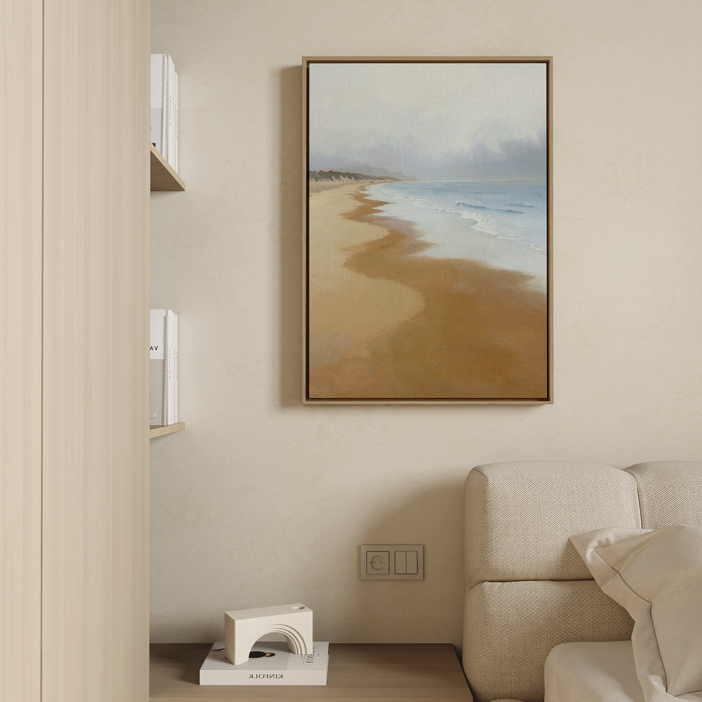 The Endless Shoreline Canvas Art - Vertical, featuring a sandy beach and ocean, hangs on a beige wall above a light-colored couch. Nearby, books and a decorative object on a wooden shelf enhance the calm, minimalistic ambiance.