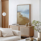 A minimal modern living room features a wooden sideboard topped with vases, books, and the Endless Shoreline Canvas Art - Vertical of a serene beach. A white armchair and small potted plant in the foreground enhance the tranquil vibe.