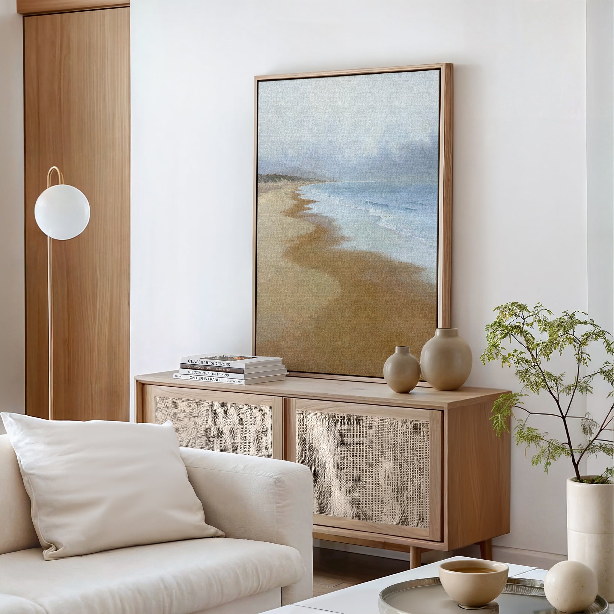 A minimal modern living room features a wooden sideboard topped with vases, books, and the Endless Shoreline Canvas Art - Vertical of a serene beach. A white armchair and small potted plant in the foreground enhance the tranquil vibe.