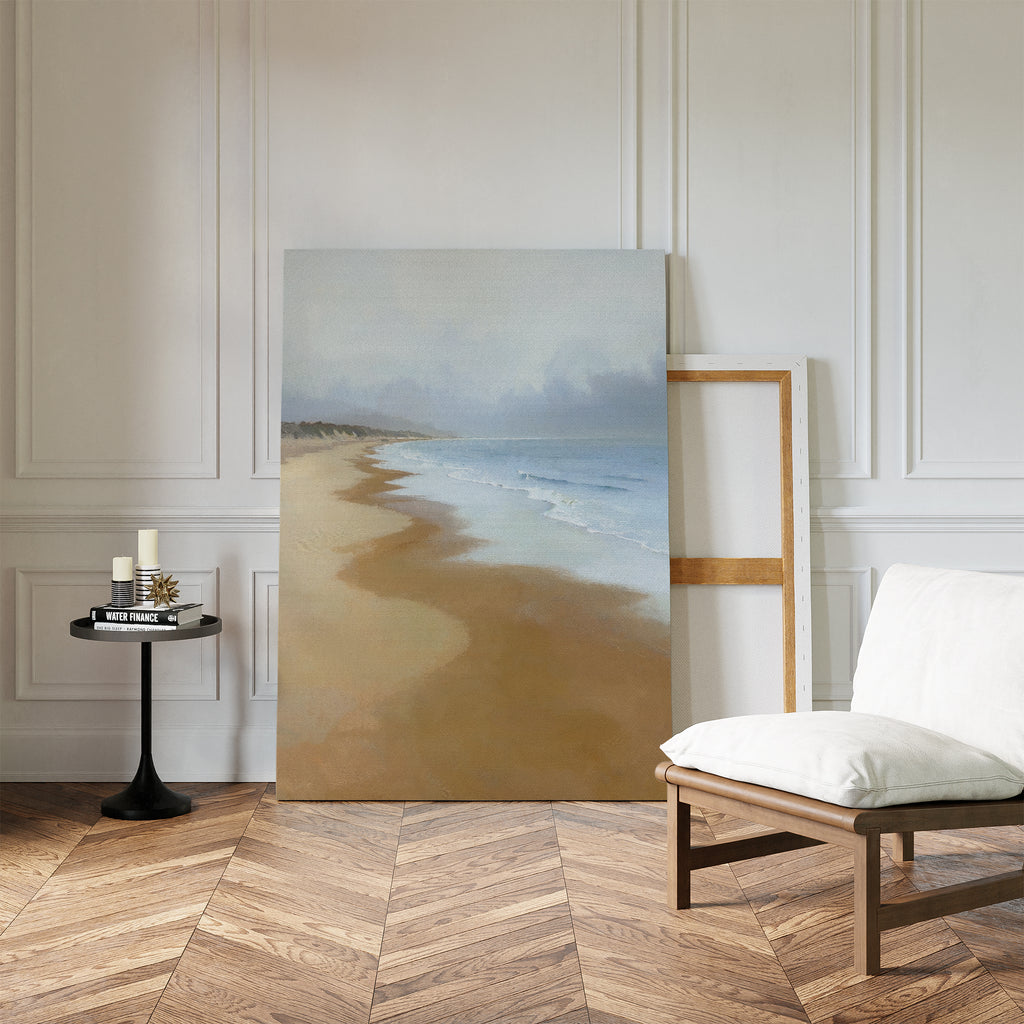 The Endless Shoreline Canvas Art - Vertical leans against a white paneled wall beside an empty wooden frame. Nearby, a white cushioned chair and round table with candles and books sit on the herringbone wood floor.