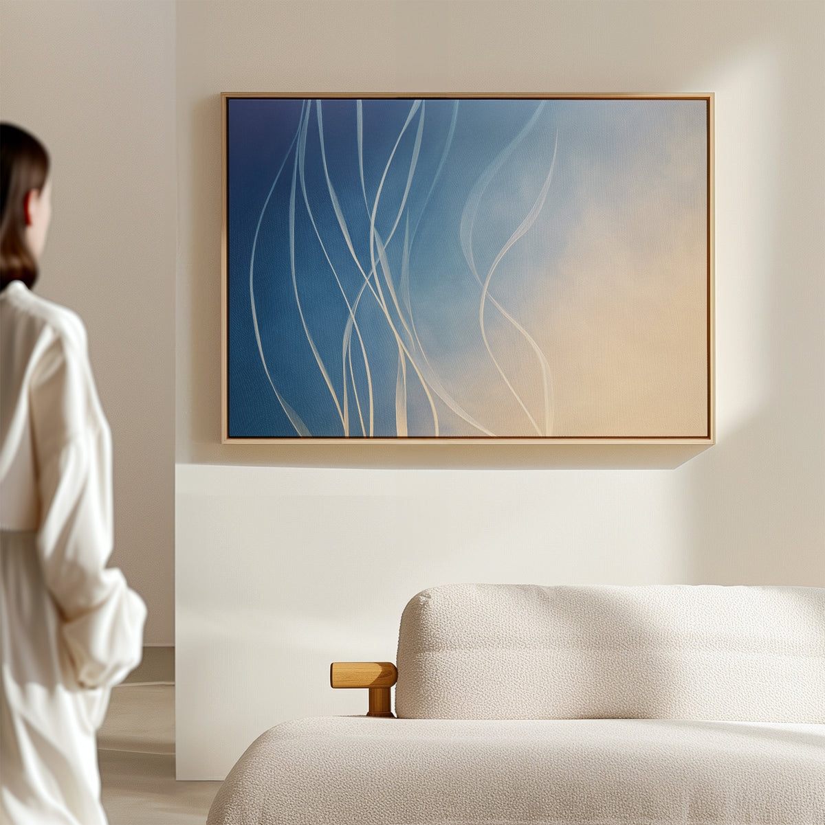 A person in a white shirt stands in a minimal, bright room, admiring the Ethereal Lines Canvas Art - Horizontal—an abstract piece with blue, beige, and white curves—adding an elegant modern touch above the beige sofa.