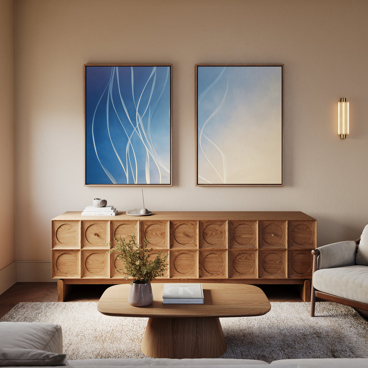 A modern living room features a wooden sideboard, coffee table, beige armchair, small plant, and the Ethereal Lines - Set Of 2 blue and beige abstract prints above the sideboard. Warm lighting adds a cozy touch of modern art.