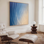 A modern living room with a white leather lounge chair and ottoman, a small wooden side table with a white lamp, and the Ethereal Lines Canvas Art - Square featuring blue and beige tones. Light streams through a tall window.