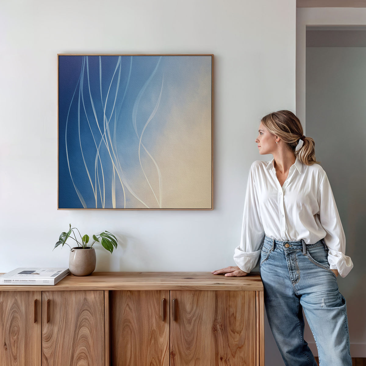 A woman in a white blouse and jeans stands beside a wooden cabinet with a plant and books, admiring the Ethereal Lines Canvas Art - Square, an abstract blue and beige print displayed on the living room wall.