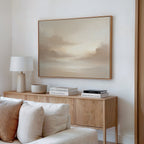 A cozy living room features a light wood sideboard, beige sofa, and stacked books. Above, the Ethereal Veil Canvas Art - Horizontal hangs on the white wall, its soft cloud tones adding tranquility to the space.