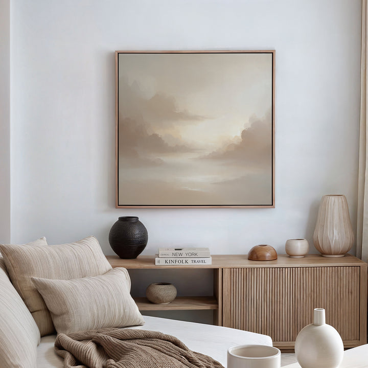 A minimalist living room with neutral tones includes a beige sofa, wooden sideboard with vases and books, and the Ethereal Veil Canvas Art - Square displaying an abstract landscape. Soft natural light brightens the space.