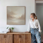 A woman in a white blouse and jeans leans against a wooden console table, admiring the Ethereal Veil Canvas Art - Square. A potted plant and books on the table complete the calm, minimalist setting.