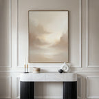A minimalist interior features a marble console table, decor accents, and the Ethereal Veil Canvas Art - Vertical: a serene beige cloudy sky print elegantly displayed on a paneled wall.