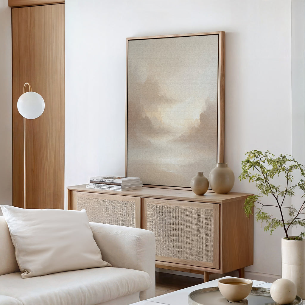A minimal, modern living room features a cream sofa with a cushion, a wooden sideboard with ceramic vases and books, the Ethereal Veil Canvas Art - Vertical as serene wall decor, a potted plant, and a round hanging light.