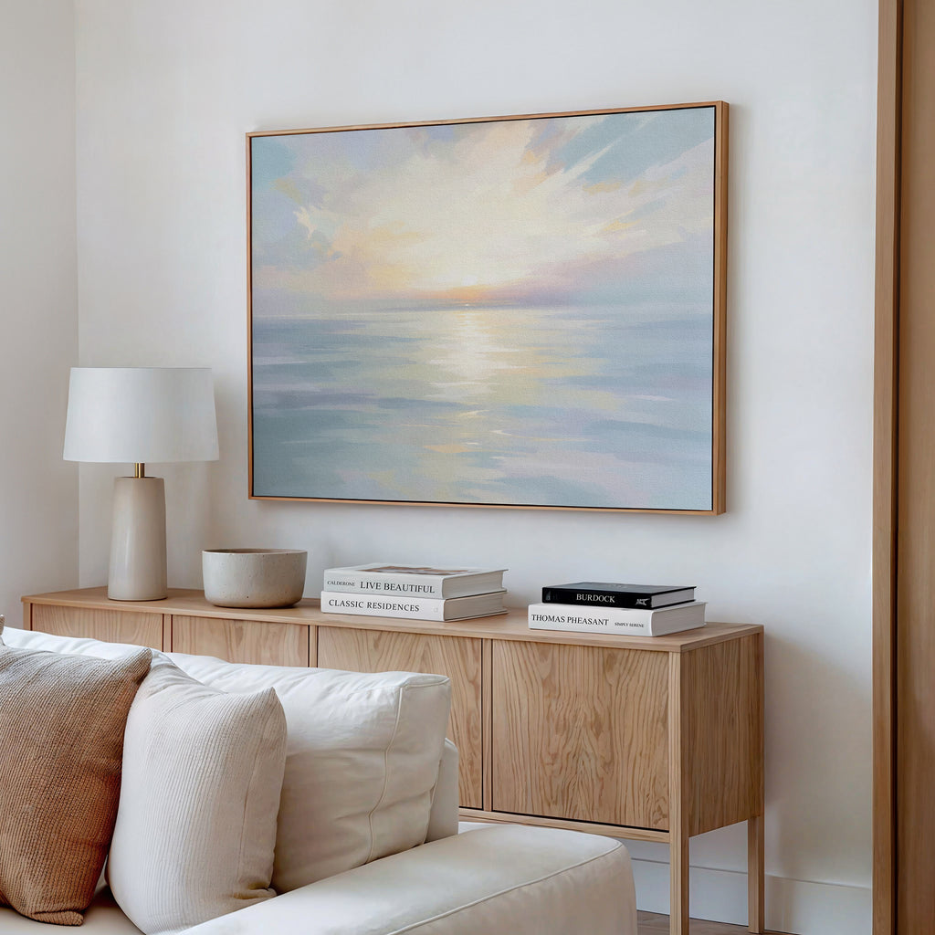 A serene living room showcases the Ethereal Waters Canvas Art - Horizontal, depicting a glowing sky above calm seas, along with a wooden sideboard, white lamp, stacked books, and cream sofa cushions for a tranquil vibe.