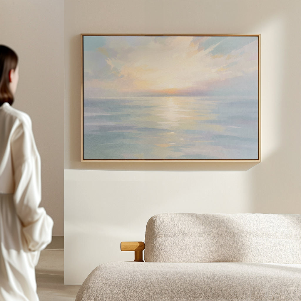 A person stands in a softly lit room, admiring the "Ethereal Waters Canvas Art - Horizontal," a serene pastel seascape print with a subtle sunset, elegantly framed and displayed above a light-colored sofa.