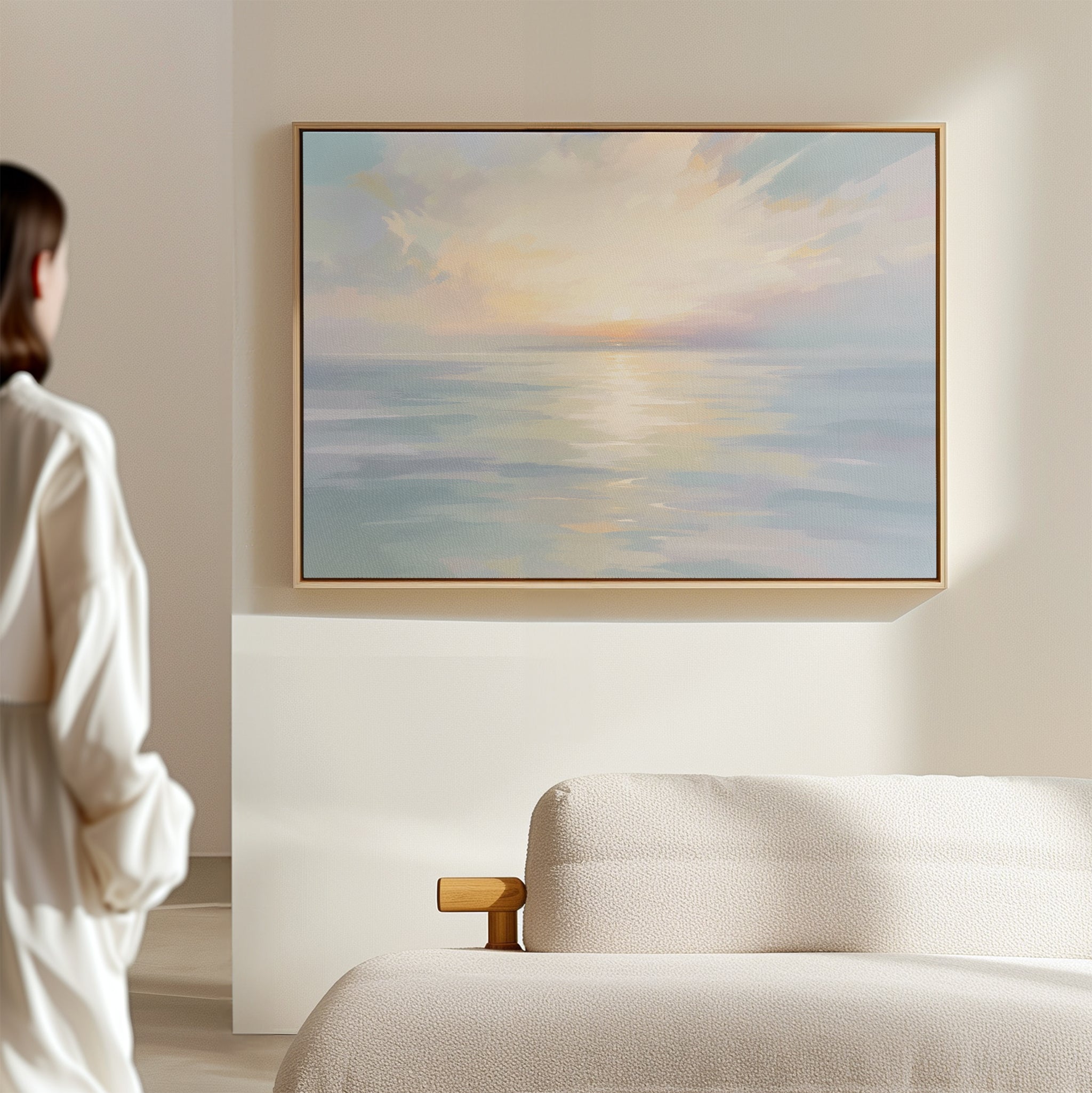A person stands in a softly lit room, admiring the "Ethereal Waters Canvas Art - Horizontal," a serene pastel seascape print with a subtle sunset, elegantly framed and displayed above a light-colored sofa.