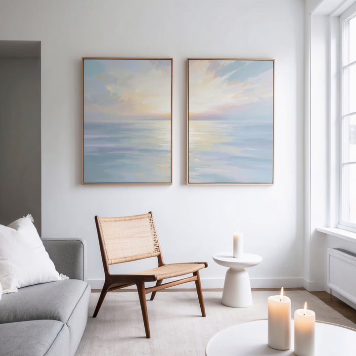 A modern living room features a gray sofa, woven chair, white side table with a lit candle, and "Ethereal Waters - Set of 2" coastal seascape paintings on the wall. Three glowing candles create a cozy, serene atmosphere.