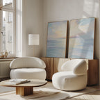A modern living room with two curved white chairs, a round wooden coffee table, a floor lamp, and the Ethereal Waters - Set of 2 wall art displayed on a wooden sideboard by a large window filled with natural light.