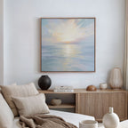 A minimalist living room with a beige sofa, neutral pillows, a wooden sideboard topped with vases and books, and “Ethereal Waters Canvas Art - Square”—a serene ocean scene—framed on the white wall.