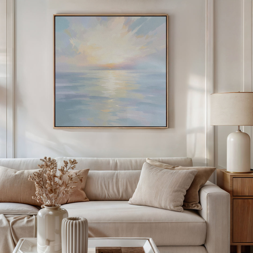 A cozy living room with a beige sofa, cream pillows, a small bouquet in a vase, a table lamp, and the Ethereal Waters Canvas Art - Square featuring a pastel seascape with the sun above the water hanging on the wall.
