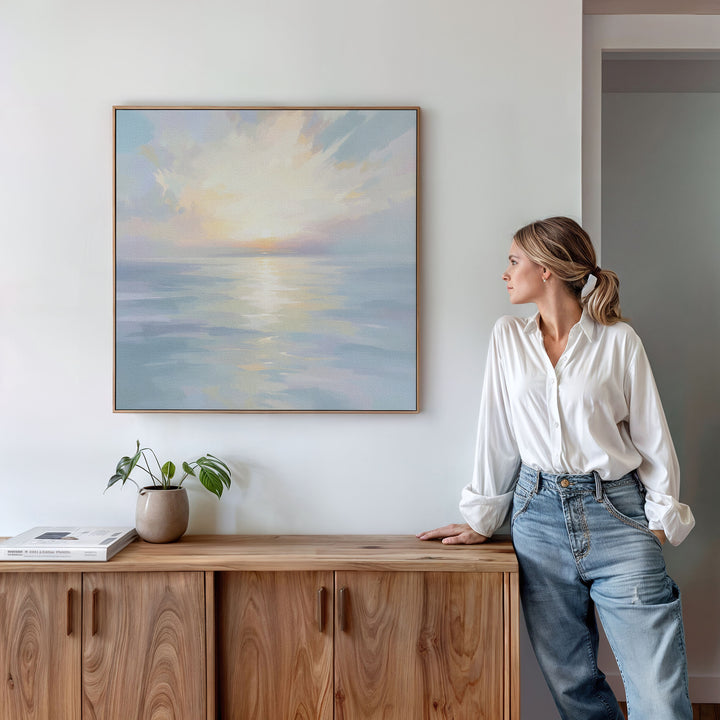 A woman in a white blouse and jeans stands by a wooden cabinet, admiring the Ethereal Waters Canvas Art - Square, which depicts a serene sea at sunrise. A small plant and books decorate the cabinet beneath the tranquil wall art.