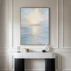 The Ethereal Waters Canvas Art - Vertical, featuring a tranquil sunrise or sunset over still water, is displayed in a frame on a beige wall above a white marble console table with black legs, accented by candles and minimalist vases.