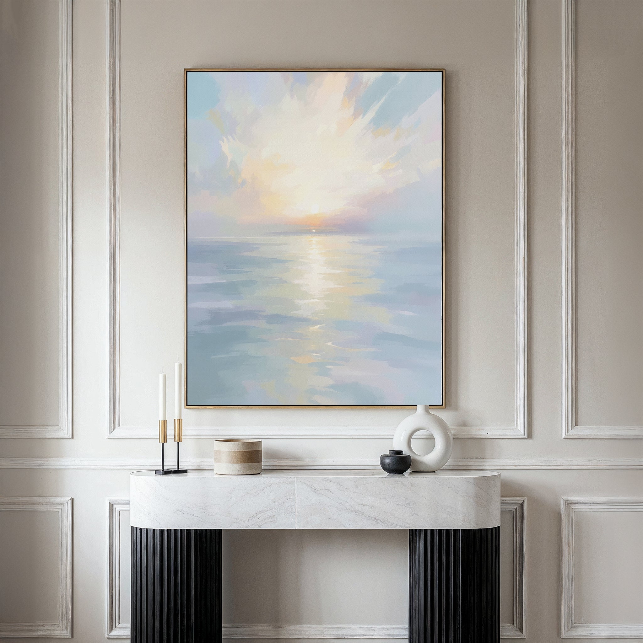 The Ethereal Waters Canvas Art - Vertical, featuring a tranquil sunrise or sunset over still water, is displayed in a frame on a beige wall above a white marble console table with black legs, accented by candles and minimalist vases.