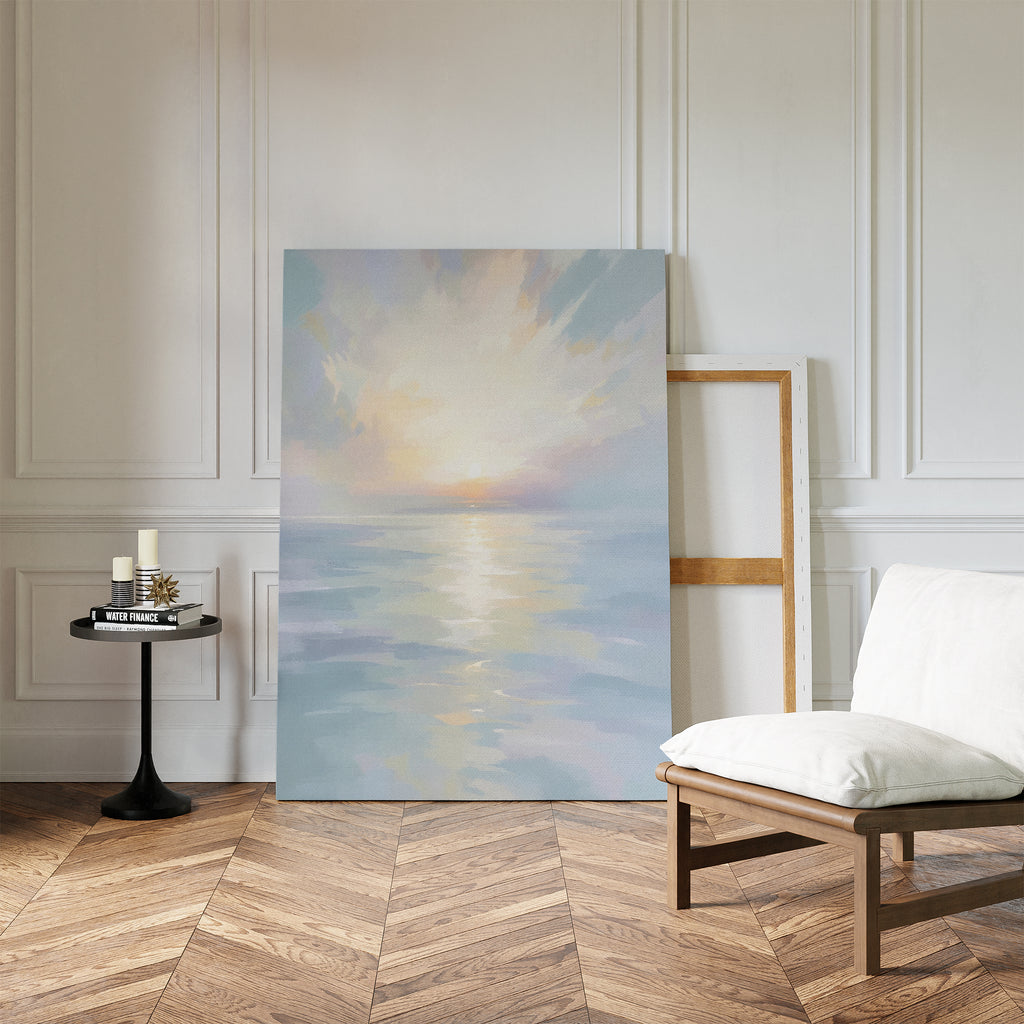 The Ethereal Waters Canvas Art - Vertical, depicting a serene sunrise over calm water, leans against a white-paneled wall in a stylish room next to an empty frame, cushioned wooden chair, and small round table with candles and books.