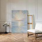 The Ethereal Waters Canvas Art - Vertical, depicting a serene sunrise over calm water, leans against a white-paneled wall in a stylish room next to an empty frame, cushioned wooden chair, and small round table with candles and books.