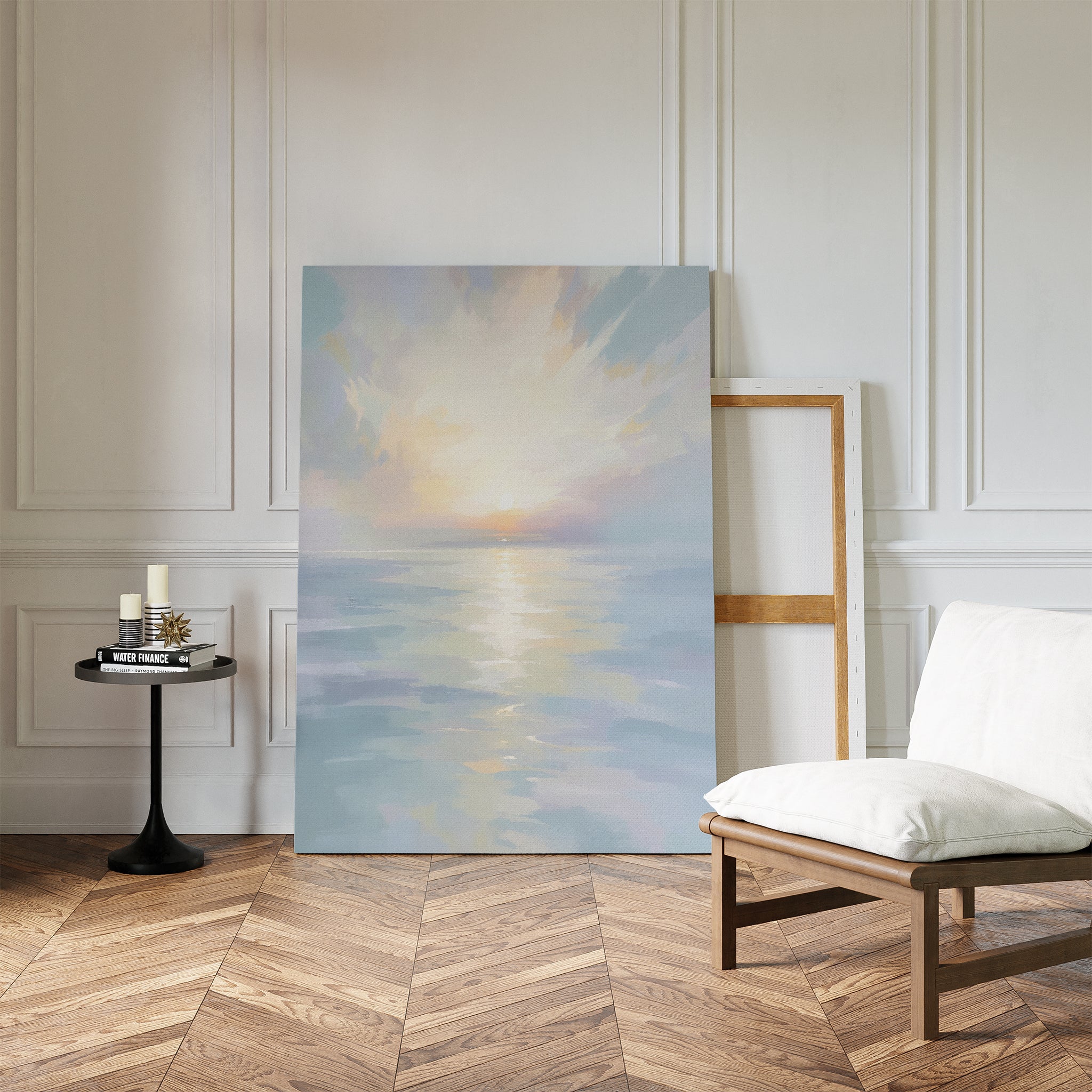 The Ethereal Waters Canvas Art - Vertical, depicting a serene sunrise over calm water, leans against a white-paneled wall in a stylish room next to an empty frame, cushioned wooden chair, and small round table with candles and books.