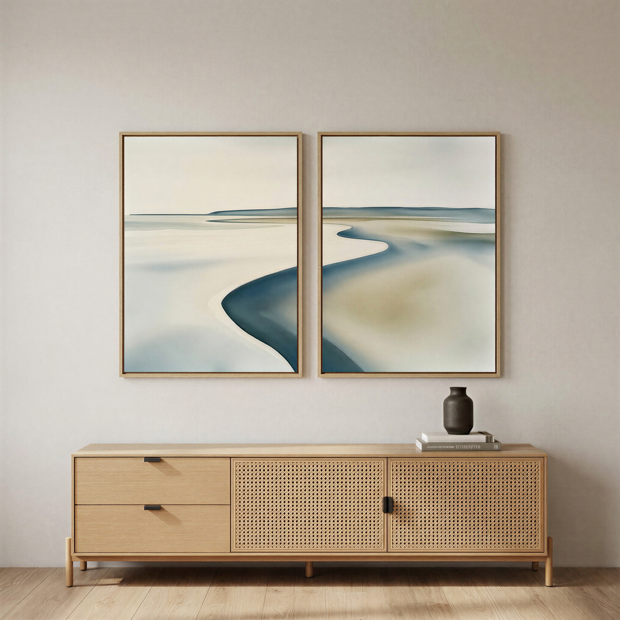 A minimalist room features a light wood sideboard, black vase, and books. Above, the Serene Coastline Set of 2 coastal wall art in soft blue and beige hangs on a light wall with wooden floors—ideal for modern home decor lovers.