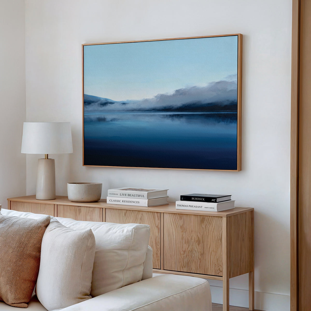 A cozy living room with a beige couch, wooden sideboard, and books is enhanced by the Fjord Mist Canvas Art - Horizontal, a large framed landscape of a misty lake and mountains above the sideboard for tranquil Nordic decor.