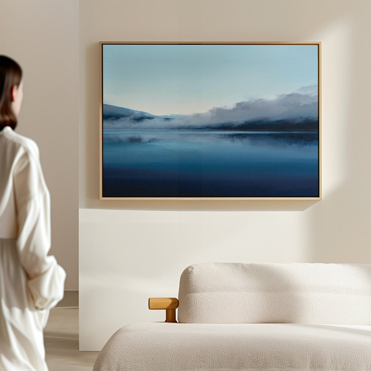 A woman stands in a minimal, neutral-toned room, admiring the Fjord Mist Canvas Art - Horizontal—a serene landscape of a calm blue lake and misty mountains—elegantly displayed as Nordic wall art above a beige couch.