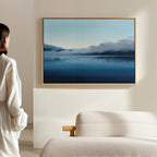 A woman stands in a minimal, neutral-toned room, admiring the Fjord Mist Canvas Art - Horizontal—a serene landscape of a calm blue lake and misty mountains—elegantly displayed as Nordic wall art above a beige couch.