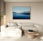 Minimalist living room with a light beige sectional sofa, wooden coffee table, and the Fjord Mist Canvas Art - Horizontal depicting serene blue mountains and water. A tall plant and sheer curtains complete this tranquil decor.