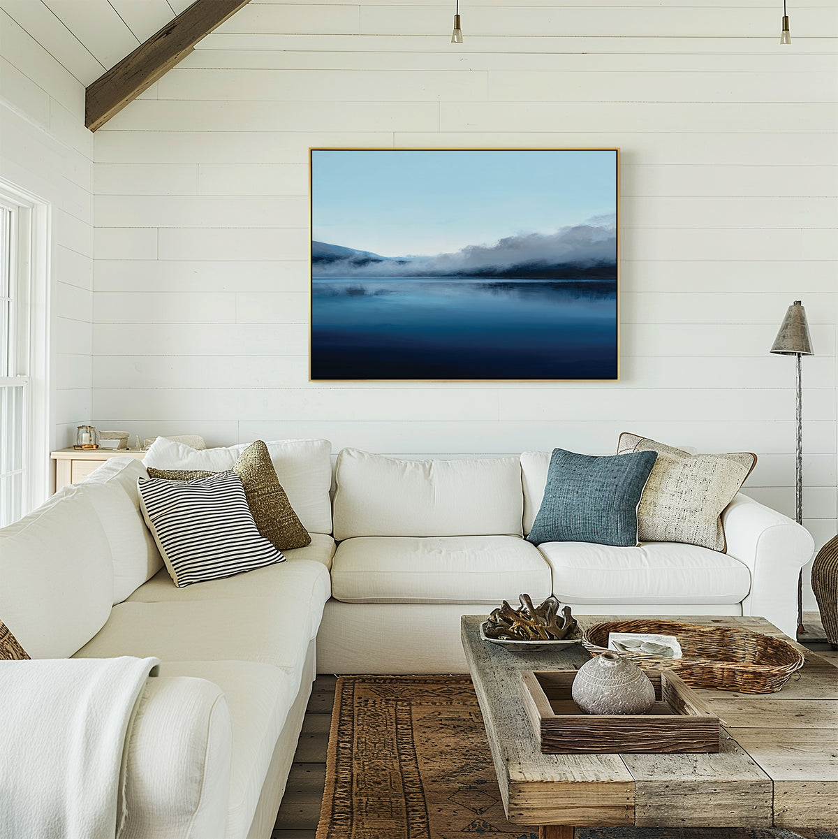 A cozy, modern living room with white shiplap walls, a white sectional with pillows, a wooden coffee table, and the Fjord Mist Canvas Art - Horizontal—a large landscape of misty blue mountains—hung above the couch.