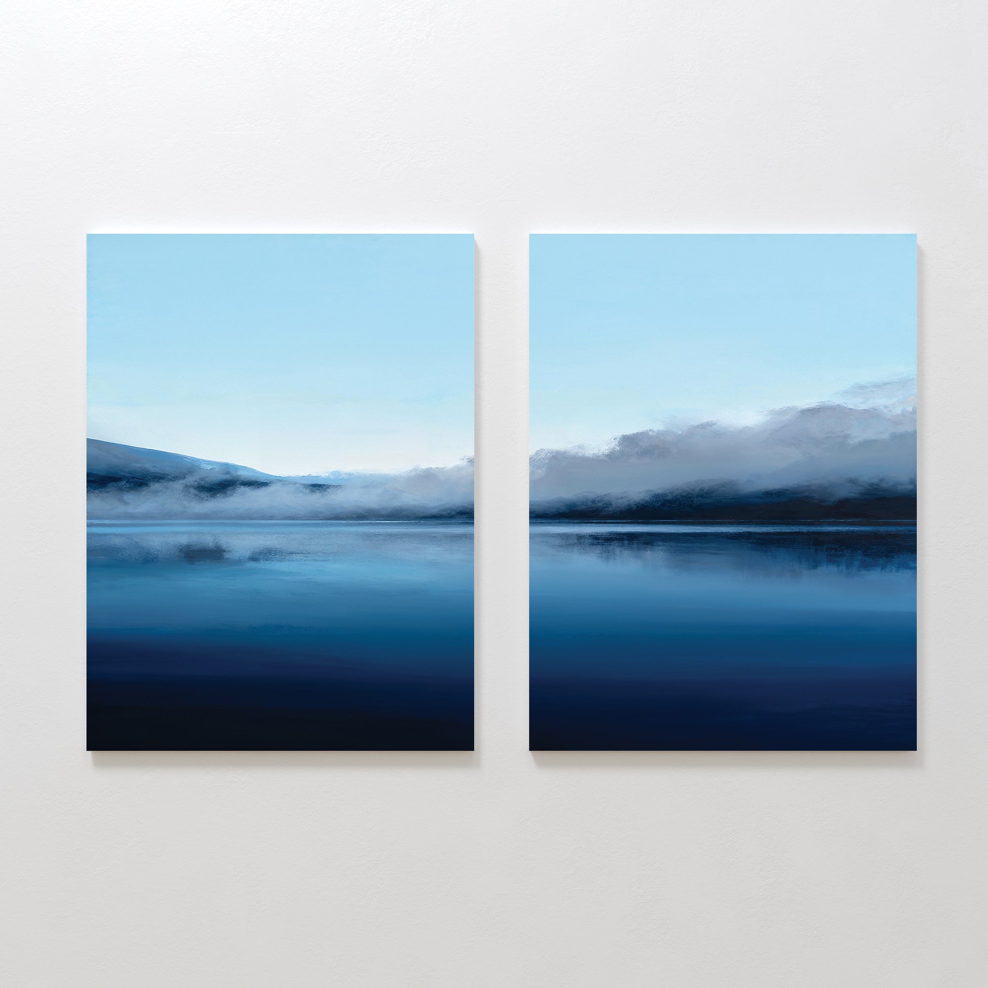 The Fjord Mist - Set Of 2 features canvas paintings of a blue lake with misty mountains and fog beneath a clear sky, hung side by side on a white wall. The calm water beautifully reflects the tranquil landscape.