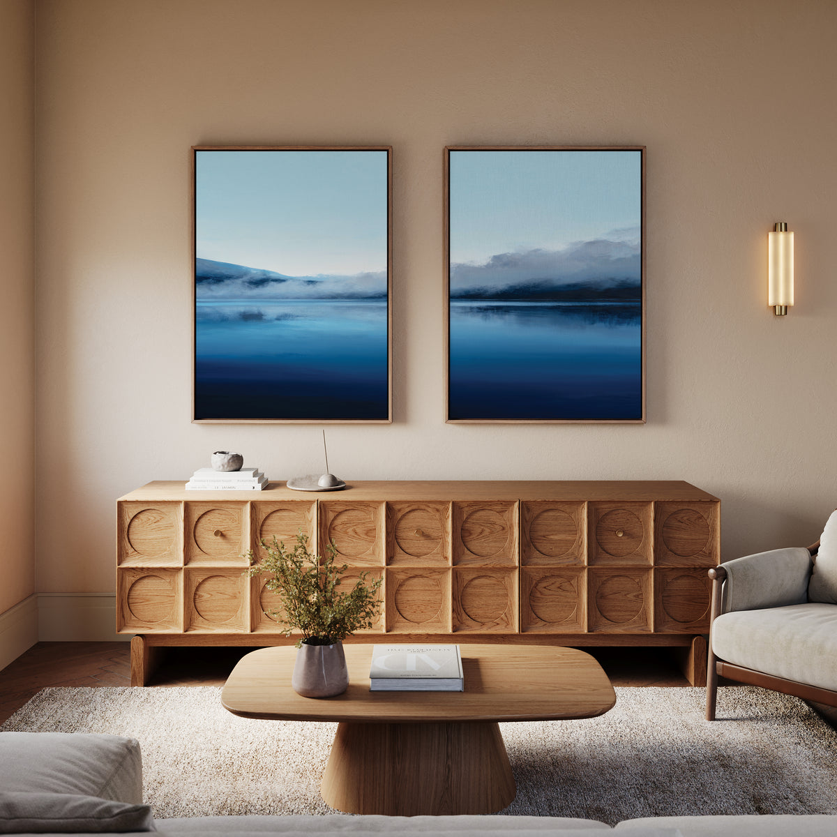 A cozy living room with a wooden sideboard, coffee table, beige walls, light sofa, and the Fjord Mist - Set Of 2 landscape prints above the sideboard. A wall sconce offers soft light and a vase with greenery decorates the table.