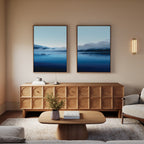 A cozy living room with a wooden sideboard, coffee table, beige walls, light sofa, and the Fjord Mist - Set Of 2 landscape prints above the sideboard. A wall sconce offers soft light and a vase with greenery decorates the table.