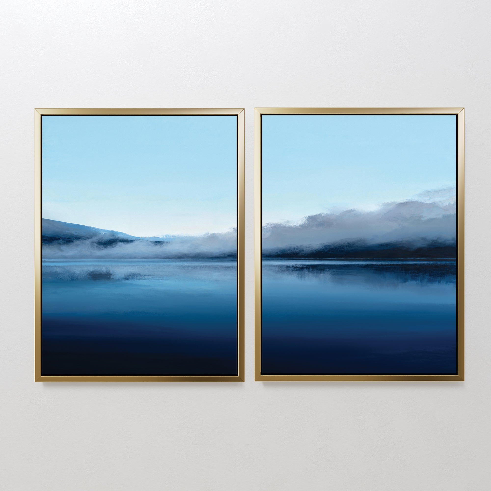 Fjord Mist - Set Of 2 features framed serene landscape prints side by side, each depicting a calm blue lake with misty hills and light blue sky. The minimalist blue waters artwork is set in gold frames against a plain white wall.