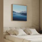 A neatly made bed with white pillows and bedding sits beneath the Fjord Mist Canvas Art - Square, featuring a serene misty lake and mountains, adding tranquility to the minimalist bedroom decor. A bare branch decorates a nearby table.