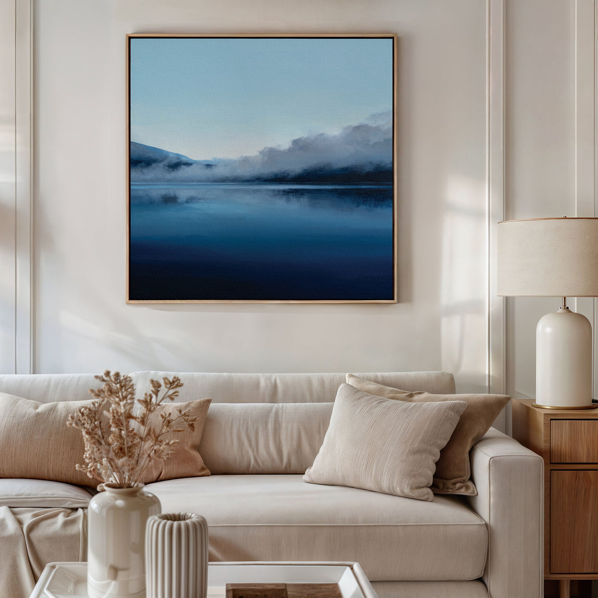 A modern living room with a beige sofa, neutral cushions, a white lamp, wooden side table, and the Fjord Mist Canvas Art - Square above the sofa. A vase with dried flowers sits on the coffee table.