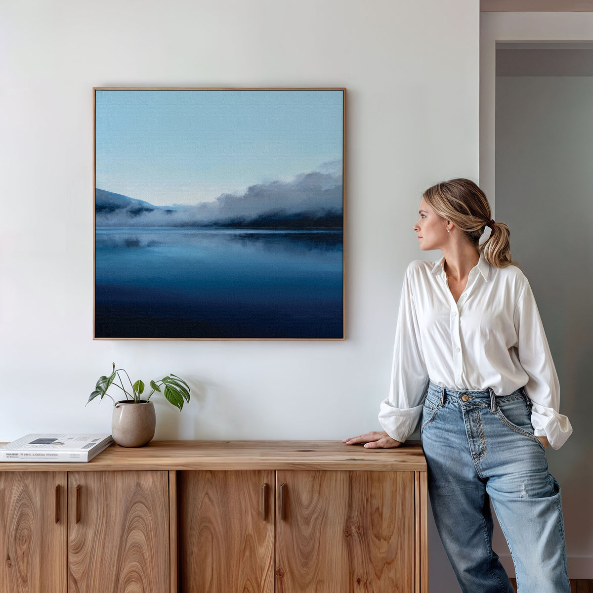 A woman in a white blouse and jeans stands by a wooden sideboard, admiring the Fjord Mist Canvas Art - Square, featuring a misty lake and mountains. A small plant and book add to the tranquil bedroom decor on the sideboard.